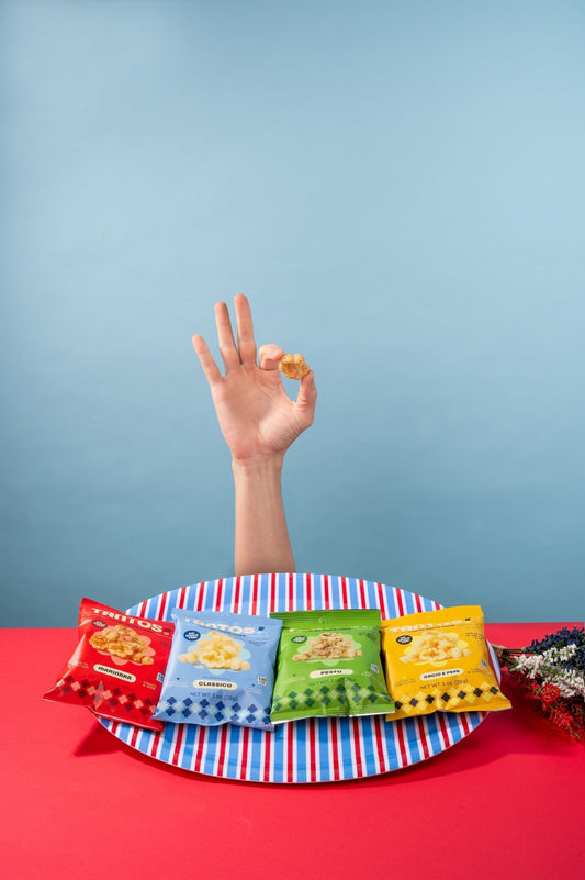 A hand giving an OK sign holding a Tantos puffed pasta chip above a striped plate with four Tantos flavor bags (Marinara, Classico, Pesto, Cacio e Pepe) on a red table