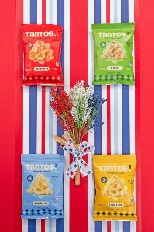 Four flavors of Tantos puffed pasta chips (Marinara, Pesto, Classico, Cacio e Pepe) arranged around a patriotic red, white, and blue floral bouquet on striped background