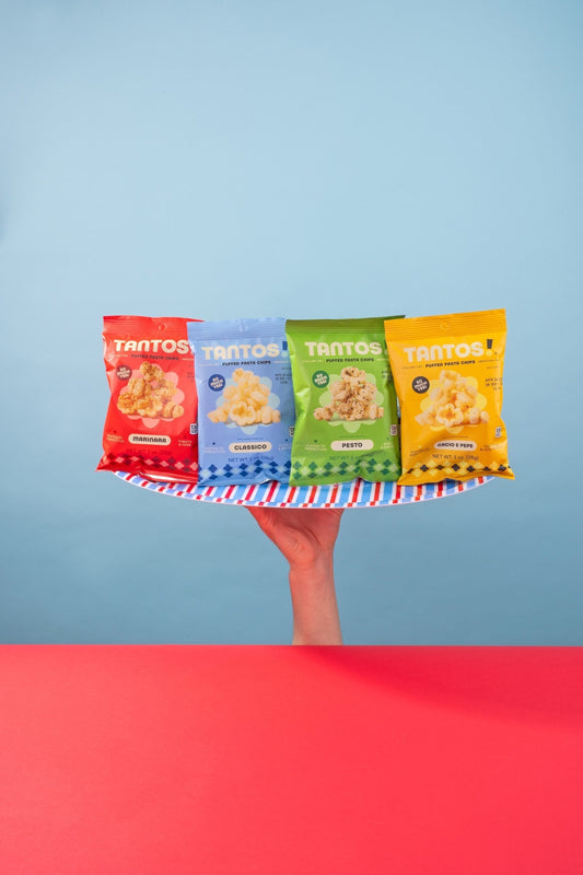 A hand holds up a striped plate displaying four Tantos puffed pasta chips bags in Marinara, Classico, Pesto, and Cacio e Pepe flavors against a blue background with a red surface below.