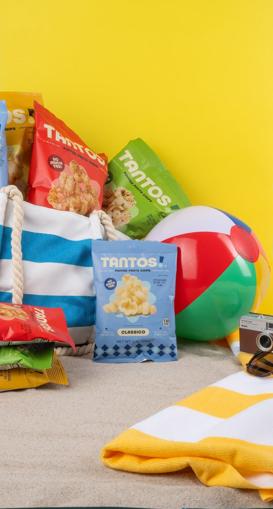 Beach-themed scene with Tantos puffed pasta chips bags (Classico and other flavors) spilling from a striped tote bag, surrounded by a beach ball, camera, sunglasses, and towel on sand