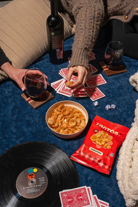 Overhead view of a game night scene with Tantos Marinara puffed pasta chips bag, a bowl of chips, wine glasses, playing cards, dice, and a vinyl record on a blue blanket