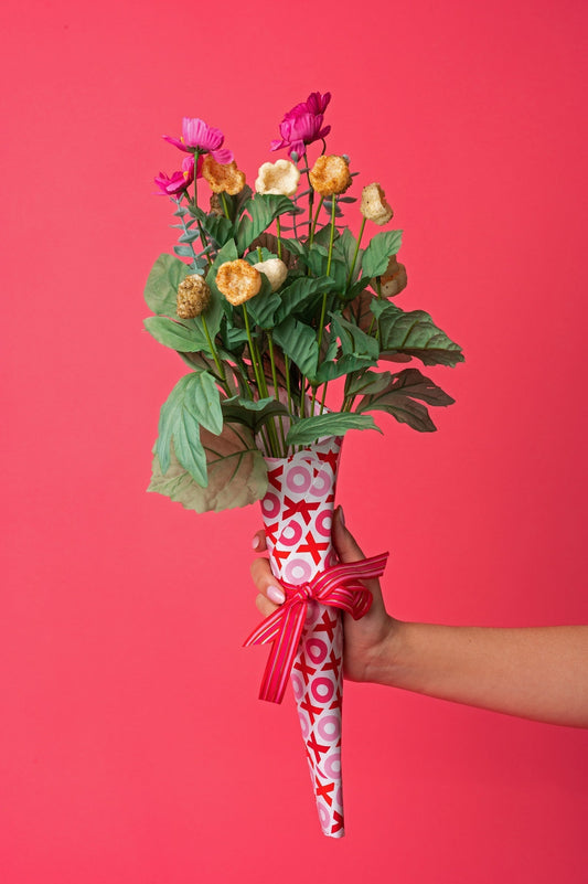 A hand holds a bouquet wrapped in red and white XOXO paper, with pink flowers and Tantos puffed pasta chips styled among the greenery against a coral background