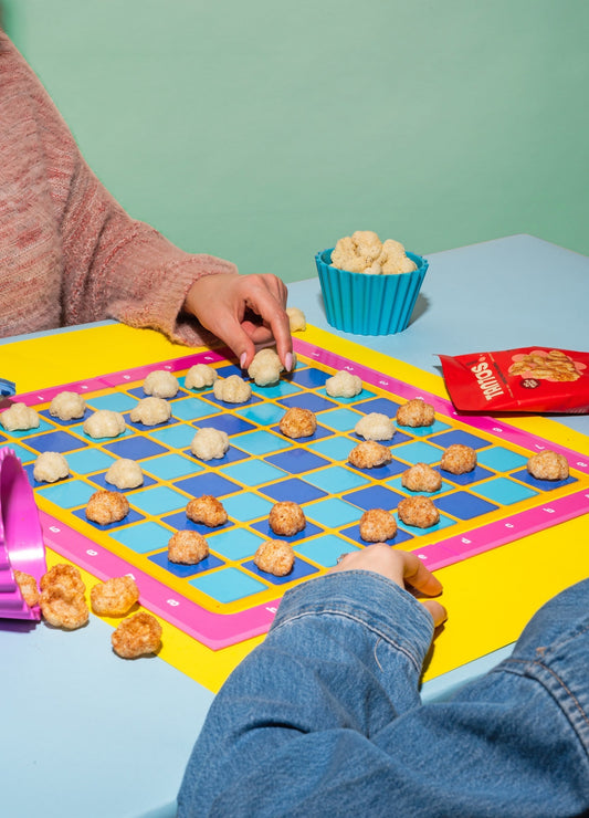 Two people playing checkers on a colorful board using Tantos puffed pasta chips as game pieces, with a bowl of chips and a red Tantos bag nearby