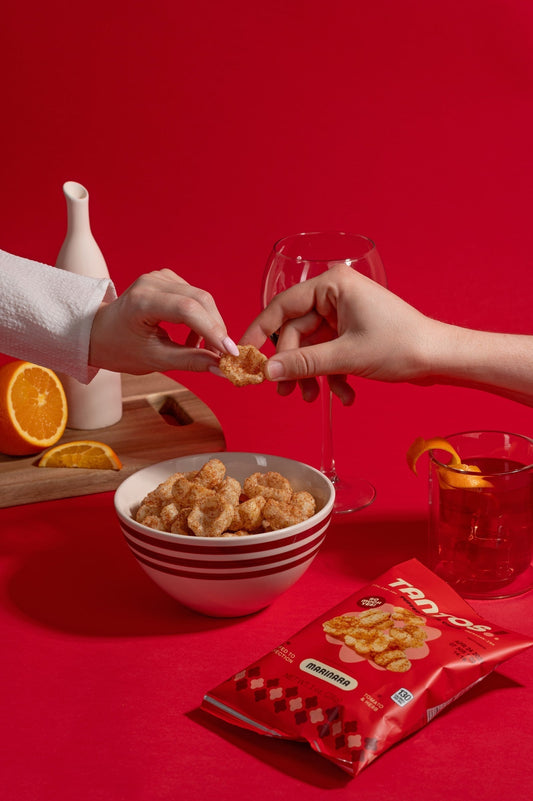 Two hands share a Tantos Marinara puffed pasta chip over a bowl of chips, wine glass, and orange garnish on a red background with the Tantos bag in front